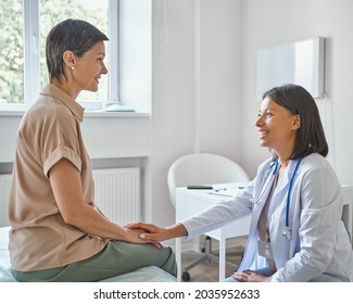 Medical ethics. Young smiling caring african female doctor supporting and comforting happy middle-aged woman patient in hospital, holding hand and expressing care during consultation in medical center