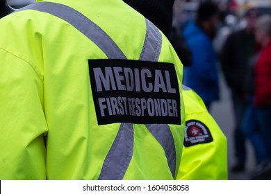 Medical First Responder Wearing A Bright Yellow Coat With A Grey Reflective Cross. The Person Is Back On To The Camera. The Emergency Response Person Is Part Of The St. John's Ambulance Organization. 