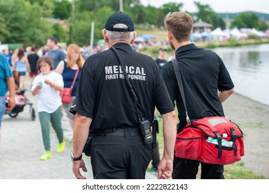 Medical first responders walk along a road wearing black uniforms, with the medical first responder in grey letters across the back of the paramedic. The EMT is carrying a red first aid kit and radio. - Powered by Shutterstock