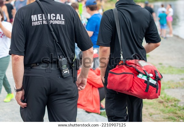 Medical first responders walking along a road\
wearing black uniforms, with medical first responder in grey\
letters across the back of the paramedic. The EMT is carrying a red\
first aid kit and\
radio.