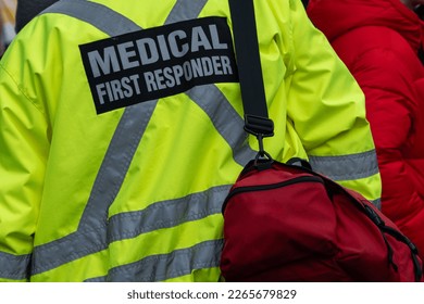 Medical first responders walking in a street among a crowd of people. The EMTs are carrying a red first aid bag. The emergency personnel has bright yellow reflective jackets with grey stripes. - Powered by Shutterstock