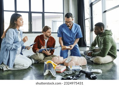 medical instructor showing wound care simulator to multiethnic team with clipboards near CPR manikin and medical equipment in training room, effective life-saving skills concept