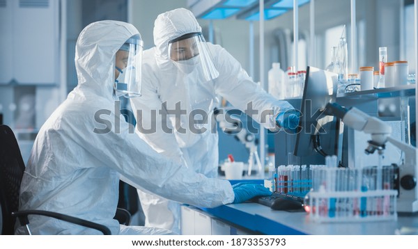Medical Laboratory: Team of Microbiology\
Scientists Wearing Sterile Coveralls, Face Shields and Masks Talk,\
Use Computer to Analyse Test Tube Blood Samples and Develop\
Vaccine, Drugs and\
Antibiotics