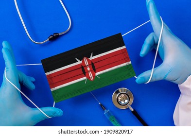 Medical Mask With The Flag Of Kenya In The Hands Of A Doctor And Stethoscope, Flat Lay. Coronavirus Pandemic