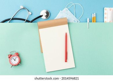 Medical Mask With Stethoscope And Blister Of Pills On A Blue Background Top View. Medical Desktop Concept. Open Notepad With Red Pen And Alarm Clock On Green. Close Up Flat Lay.