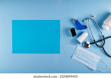 Medical mask, stethoscope, oximeter, bottle of pills and inhalers on light blue background with vibrant blue rectangular sheet of paper for text placement. Asthmatic breathing symptoms Concept