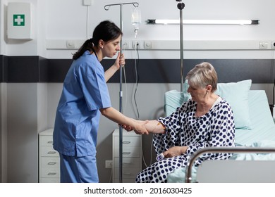 Medical Nurse Helping Senior Woman Patient Getting Up From Bed In Hospital Room, With Iv Drip Bag Attached While She Gets Intravenous Treatment And Oxymeter Measuring Blood Oxygen Saturation.