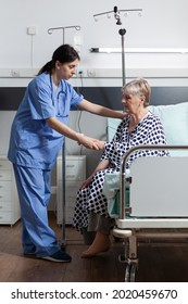 Medical Nurse Helping Senior Woman Patient Getting Up From Bed In Hospital Room, With Iv Drip Bag Attached While She Gets Intravenous Treatment And Oxymeter Measuring Blood Oxygen Saturation.