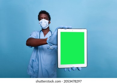 Medical Nurse Holding Vertical Green Screen On Tablet In Studio. Healthcare Assistant With Covid 19 Face Mask And Stethoscope Having Gadget With Chroma Key, Showing Isolated Mockup Template.