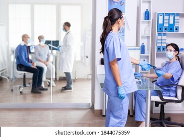 Medical Nurse Holding X-ray In Hospital Hallway During Coronavirus Outbreak Wearing Face Mask . Assistant Working On Reception Computer. Doctor Dicussing With Senior Couple