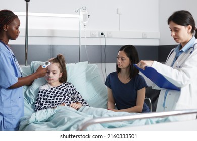 Medical Nurse Measuring Sick Little Girl Body Temperature Using Modern Thermometer While In Healthcare Pediatric Hospital Patient Recovery Ward Room. Pediatrist Writing Down Analysis Results.