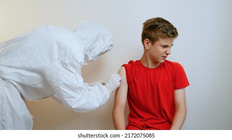 Medical Nurse In Safety Gloves And Protective Mask Is Making Vaccine Injection To Male Teen Patient In Health Clinic. Doctor Uses Hypodermic Needle And Syringe To Put Shot Of Drug As Treatment.