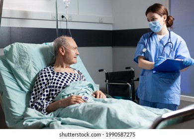 Medical Nurse In Scrubs With Chirurgical Mask Taking Notest On Cliboard During Consultation Of Sick Unwell Senior Man. Patient With Iv Drip Attached Breathing With Help From Oxygen Mask.