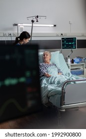 Medical Nurse In Scrubs Showing Compassion To Senior Woman Patient, Laying In Hospital Bed Breathing With Help From Oxygen Mask, Getting Intravenous Medicine From Iv Drip Bag.