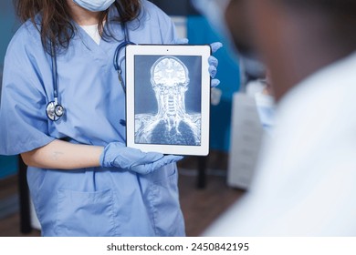 In medical office, doctor uses tablet to analyze a scan of a patient's skull. Selective focus of nurse in blue scrubs holding a device with an x-ray image. Advanced technology and expert care. - Powered by Shutterstock