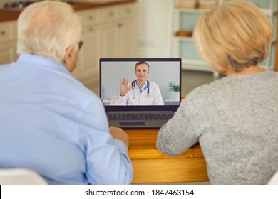 Medical Online Consultation. Back View Of A Senior Couple Sitting At A Table In Front Of A Laptop At Home And Talking To A Doctor Via Video Link. Health Care And Modern Technology For The Elderly.