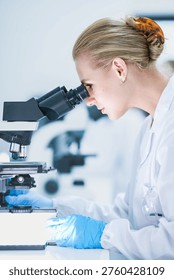 Medical pathologist looking through microscope eyepiece for cancer diagnosis. Female scientist in safety glasses analyzing biopsy slide in sterile hospital lab room.