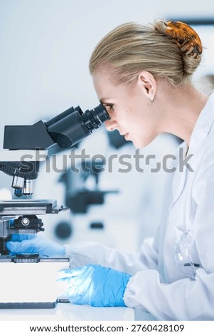 Medical pathologist looking through microscope eyepiece for cancer diagnosis. Female scientist in safety glasses analyzing biopsy slide in sterile hospital lab room.
