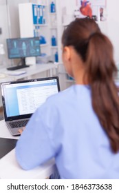 Medical Practitioner Looking At Patients Forms And Contracts Of Health Care. Healthcare Physician Using Computer In Modern Clinic Looking At Monitor, Medicine, Profession, Scrubs.