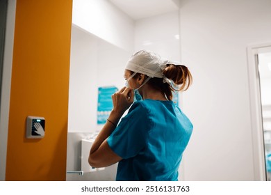 A medical professional adjusts their surgical mask in a preparation room, getting ready for an operation, highlighting the importance of hygiene and concentration in medical procedures. - Powered by Shutterstock