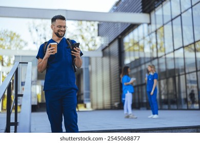 A medical professional in blue scrubs happily uses a smartphone while holding a coffee cup outside a contemporary clinic with colleagues in the background. The scene exudes positivity and teamwork. - Powered by Shutterstock