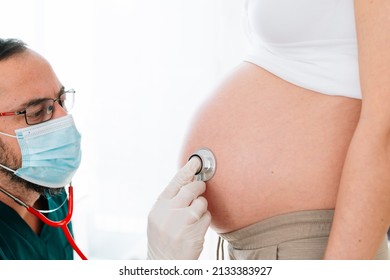 Medical Professional With A Face Mask For The Global Pandemic Of Covid 19 Listening With A Stethoscope To The Heartbeat Of A Baby In The Belly Of His Pregnant Mother. Pregnancy Health Care Concept