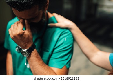 A medical professional in green scrubs, stethoscope around the neck, is distressed and being comforted by a colleague's supportive hand on the shoulder in a hospital corridor. - Powered by Shutterstock