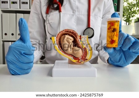 A medical professional in a lab coat and gloves holds a 3D printed model of a fetus and a bottle of pills while gesturing with a thumbs up, representing prenatal care and medication.