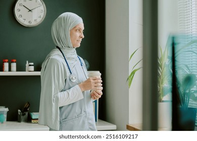 Medical professional wearing hijab standing by window holding coffee cup, with stethoscope around neck. Clock and various items visible in background on shelves - Powered by Shutterstock