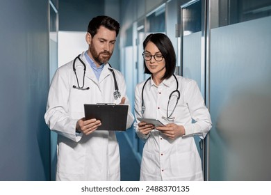 Medical professionals discussing patient records in hospital corridor. Man and woman in white coats with stethoscope and clipboard having conversation. Medical consultation and teamwork. - Powered by Shutterstock