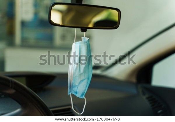 Medical protective face mask in car on the\
rearview mirror in day - Covid-19 pandemic protection on rear view\
in the vehicle - coronavirus epidemic new normal prevention\
concept