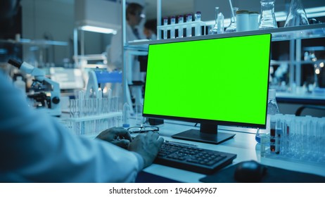 Medical Research Scientist Working On Desktop Computer With Green Screen Mock Up Template In Applied Science Research Laboratory. Lab Engineers In White Coats Conduct Experiments In The Background.