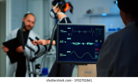 Medical researcher examining EKG image showing on monitor while patinet with mask running on cross trainer testing heart rate using electrodes.Doctor monitoring physical endurance in science sport lab