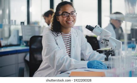 Medical Science Laboratory: Beautiful Black Scientist is Using Microscope, Looking at Camera and Smiling Charmingly. Young Biotechnology Science Specialist, Using Technologically Advanced Equipment.