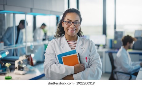 Medical Science Laboratory: Beautiful Smart Young Black Scientist Wearing White Coat And Glasses, Holds Test Books, Smiles Looking At Camera. Diverse Team Of Specialists. Medium Portrait Shot