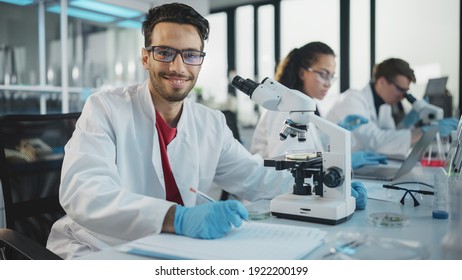 Medical Science Laboratory: Handsome Latin Scientist Analysing Samples and Uses Microscope, Looking at Camera and Smiling Charmingly. Young Biotechnology Specialist, Using Advanced Equipment. - Powered by Shutterstock