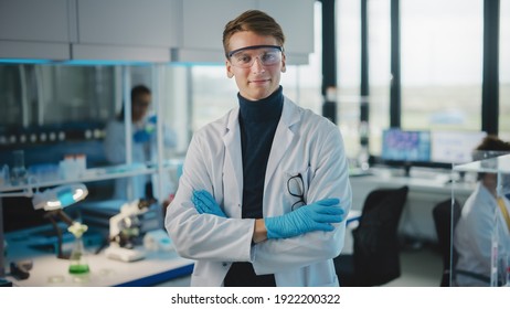 Medical Science Laboratory: Handsome Young Scientist Wearing White Coat and Safety Glasses, Posing and Smiles Looking at Camera with Crosses Arms. Diverse Team of Specialists. Medium Portrait Shot. - Powered by Shutterstock