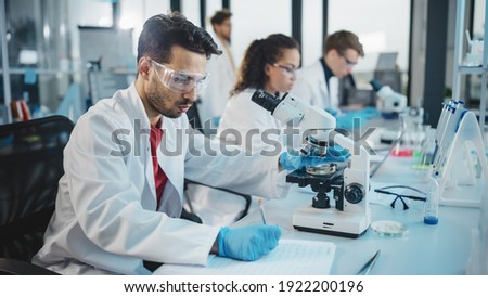 Medical Science Laboratory: Portrait Shot of a Handsome Latin Scientist Looking Under Microscope, Doing Sample Analysis. Young Biotechnology Specialist, Using Advanced Equipment.