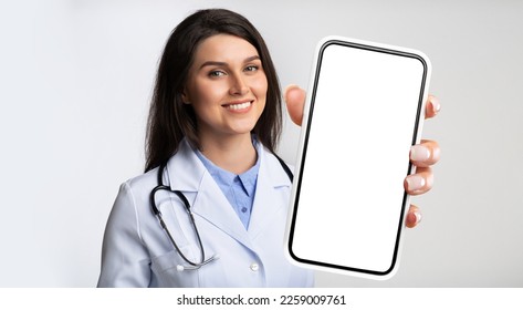 Medical Services. Smiling Female Doctor Holding Smartphone With Blank Screen In Her Hand, Beautiful Therapist Woman In Uniform Showing Empty Cellphone, Recommending Mobile App Or Website, Mockup - Powered by Shutterstock