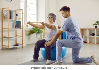 Medical specialist at modern rehabilitation center helping senior patient do physiotherapy exercises on fit ball. Old lady with osteoporosis sitting on fitball, holding rubber band and doing exercises - Powered by Shutterstock