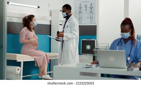 Medical Specialist And Pregnant Woman With Face Mask At Checkup Visit Talking About Bottle Of Pills. Obstetrician Using Medicine To Help Patient With Pregnancy Belly, Giving Maternity Advice.