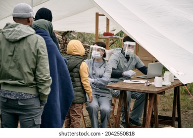 Medical specialists in protective masks sitting at table in tent and checking health of migrants standing in line - Shutterstock ID 2042550818
