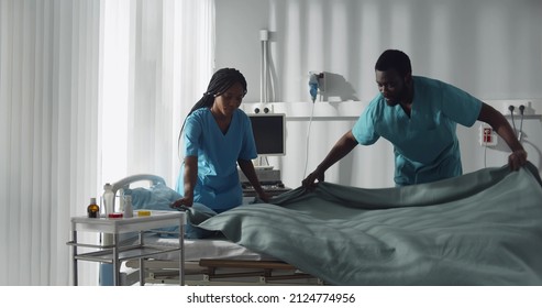 Medical Staff Cleaning Up Room And Making Beds In Hospital. African-American Nurses Changing Sheets And Cleaning In Hospital Ward Preparing For New Patients