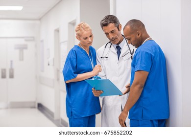 Medical Staff Having Discussion In Hospital Hallway. Mature Doctor And Multiethnic Surgeon Talking In The Corridor And Looking At Patient Files. Senior Doctor And Nurses Discussing Medical Report.