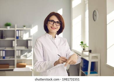 Medical Staff. Portrait Of An Experienced Female Doctor With Documents In Hands At The Clinic Office. Woman In A White Medical Gown And Glasses Looks At The Camera While Filling Out A Medical Card.