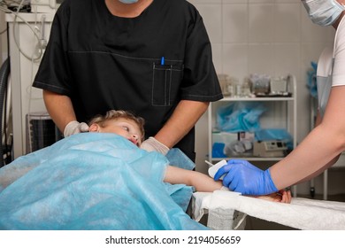 Medical Staff Prepares A Young Child For Surgery. The Baby Lies On The Operating Table, The Doctor Installs A Catheter In The Vein For The Infusion Of Medications