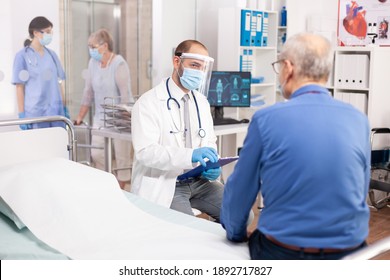 Medical staff questioning senior man in clinic consultation room wearing visor in the course of covid19. Physician practitioner in the course of private modern clinic appointment during coronavirus - Shutterstock ID 1892717827