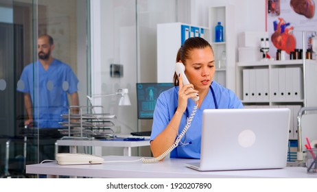 Medical Staff Talking With Patient On Phone From Hospital About Diagnosis, Male Nurse Working In Background. Healthcare Physician, Receptionist Doctor Assistant Helping With Telehealth Communication