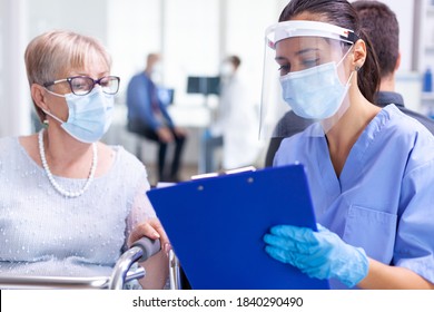 Medical staff wearing visor against coronavirus in hospital hallway talking disabled senior woman wearing face mask. Patient and medical staff in waiting area. Doctor in examination room.