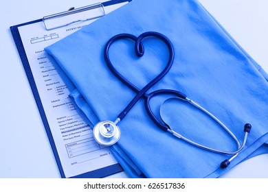 Medical stethoscope twisted in heart shape lying on patient medical history list and blue doctor uniform closeup.